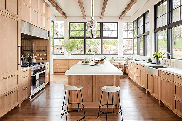 A white oak kitchen with white ceilings and medium-brown wood exposed beams. Large windows that wrap around the room are outlined in black metal and have no blinds or dressing. A deep farmhouse sink in stainless steel has a gooseneck faucet and the cook top's hood is also in stainless steel. A pair of modern white stools on black metal legs sit at the close end of the large kitchen island with a second small sink at the far end of the white concrete countertop.