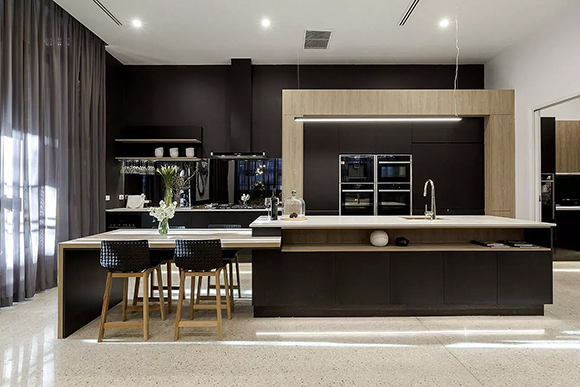 A dining area with dark curtains and light-colored wood is organized into a long, low, horizontal space with cups and silverware at the back and a long white counter with under-counter display storage at the front. A dining-height island surface has black and wood dining chairs.