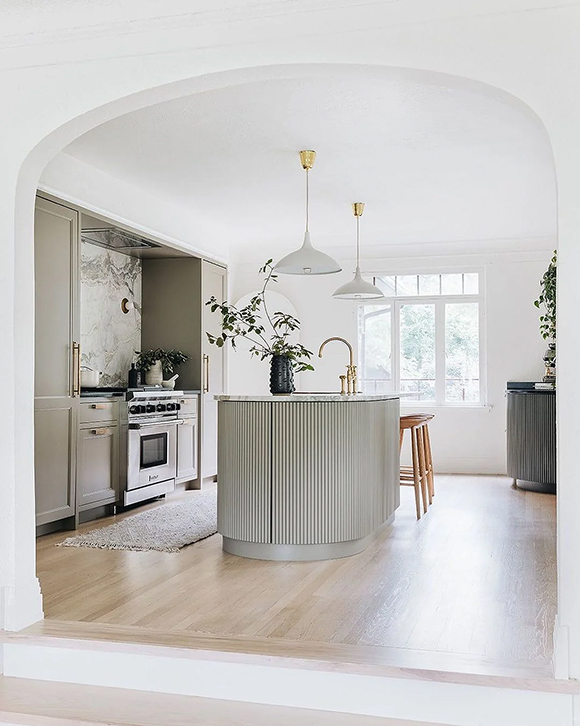 A grey pill-shaped island is the center of this grey and white kitchen with grey cabinetry and light-colored-wood flooring. Two white pendant lamps hang above the island.