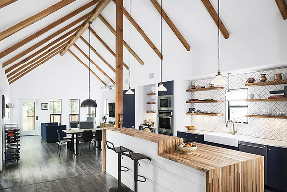 A white peaked roof with exposed wood beams is matched by a long waterfall counter that features counter height at one end and a dining height counter waterfalling off the taller surface. A pair of modern black barstools sits in front of the counter height end, while behind is a kitchen with two ovens, black cabinetry, a white farmhouse sink with gooseneck faucet, and open wood shelving on the wall behind, with herringbone white tile backsplash. Multiple pendant lights hang above the island and kitchen. A dining area is seen beyond the kitchen in an open plan space.
