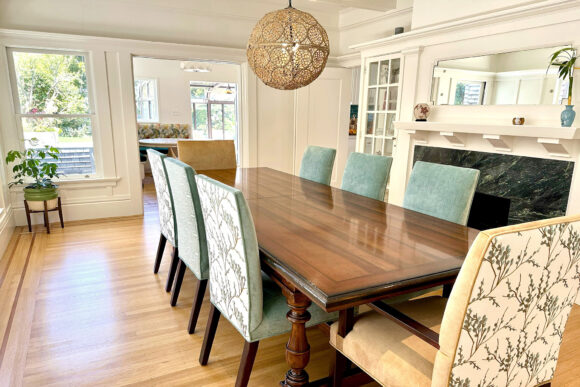 Angled view into a dining room with a long shiny rectangular dark wood dining table surrounded by 6 dining chairs in alternating chair back colors of off-white and green with a pattern that looks like the tips of tree branches or florals with only buds. A textural pierced globe pendant lamp hangs above the table. Warm cherry hardwood flooring planks run horizontally. A mirror is seen above the fireplace mantle, while the hearth is black. Green plants in stands are raised above the floor by legs.