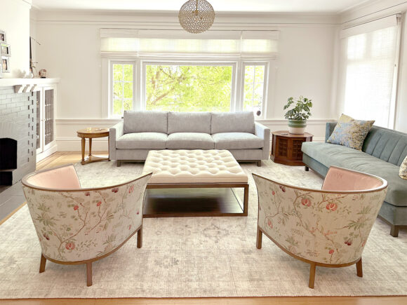 A light-colored, bright living room with pale furniture: Two tub chairs with dark wood legs, pink interior fabric and floral backs sit at the front facing away, an oversize tufted white fabric and dark wood footrest acts as a table, a blue sofa has blue and green throw pillows, while a white and light blue sofa faces you under a large window with greenery outside. A round pierced textural pendant light hangs in the center of the room. A black fireplace with white mantle is dressed with photos above and next to it is a glass cabinet of bookshelves. A green plant sits on a dark wood side table between the two sofas. Windows have roman blinds in a sheer white fabric.
