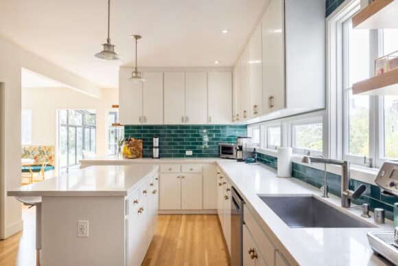 Looking down the length of an L-shaped kitchen with a matching island in the center. White quartz countertops, white cabinetry, deep green subway tiles wall finish, warm cherry hardwood flooring running longways, plenty of windows underneath the cabinets to let in natural light, a pot-filler faucet in a silver under-mount sink. 2 barn-style sing-bulb silver metal pendant lights hang above the island while recessed lights can be seen elsewhere.