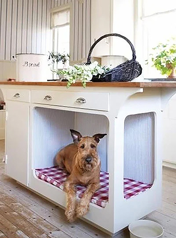An Airdale terrier lays on a red and white checked bed underneath an open sided off-white country kitchen island. On top, the counter is dressed with a basket of white flowers and a container of flour labeled "Bread". Board and batten off-white paneling can be seen just behind the island.