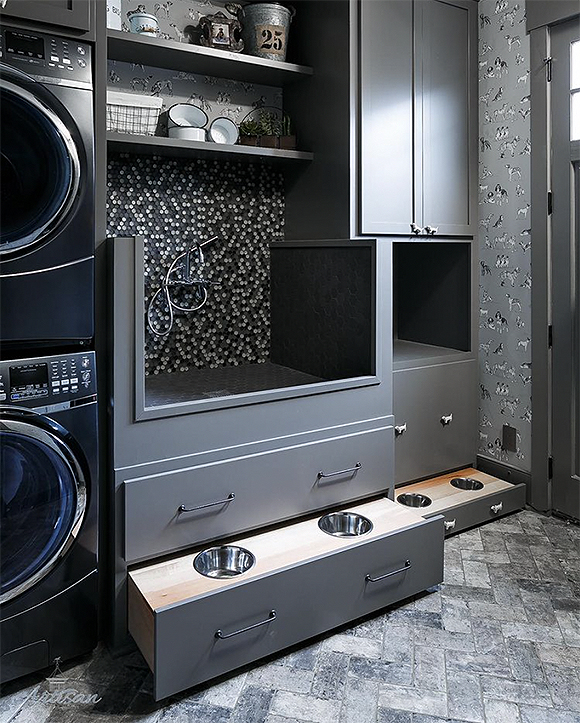 A black, silver, and white mudroom with a pair of open shelves above a tiled and rubber-finished dog-washing station. A stacked black washer and dryer set are partially seen to the left. To the right are silver cabinets, a cubby, and pull-out food and water dishes inside two front drawers. On the right, grey wallpaper has illustrations of black, white, and grey dogs. Grey trim surrounds the door frame almost out of view on the right. Herringbone grey and white tiled flooring finishes the space.