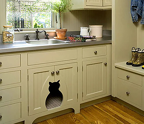 A cat-shaped cut-out sits in the center where two cabinet doors meet under a sink in a country-style kitchen. A pair of child's boots sit on a built-in seat nearby, while jars of treats and supplies sit atop the dark grey kitchen countertop above. Twisted dark metal spigots adorn the country sink below a window filled with green plants.