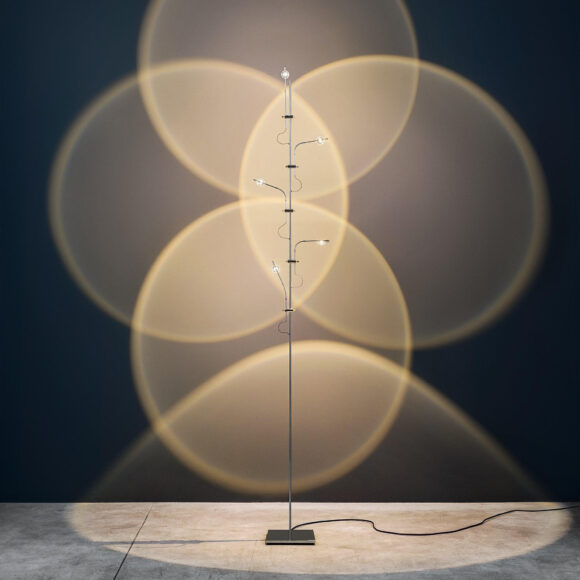 4 overlapping circles of white light are projected onto a grey wall behind. The floor lamp has a black base and silver stand with 4 adjustable LED lights.