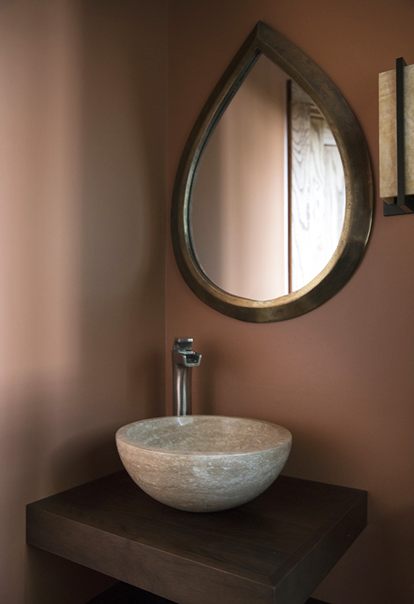 Earthy adobe-colored walls form a corner for this stone basin sink niche with a single waterfall spigot in solver, a teardrop-shaped wall-mount mirror with a hand-beaten metal frame. A wall sconce in off-white art glass and metal frame sits just at the right edge of the view. The basin sink is set atop a chunky wooden wall-mounted shelf in the corner.