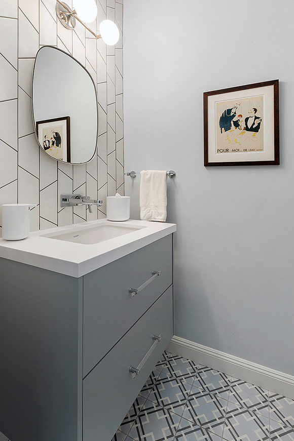A blue-grey powder room showing a portrait mirror above a single-spigot silver tap from the wall over a white one-piece quartzite counter and undermount sink. Glass and silver drawer pulls bring a touch of bling, while geometric square patterns in black and white on a field of powder grey-blue create excitement for flooring.