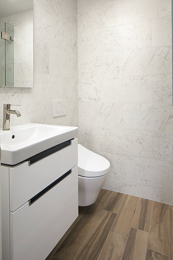 A veiny marble wall in off-white, grey, and tan counterpoints the wood-look floor tiling planks, custom white cabinet under a trough-style white ceramic sink with a brass single spigot to control water temperature. Slightly behind and partially hidden is the wall-mounted white modern toilet that appears to float above the floor.