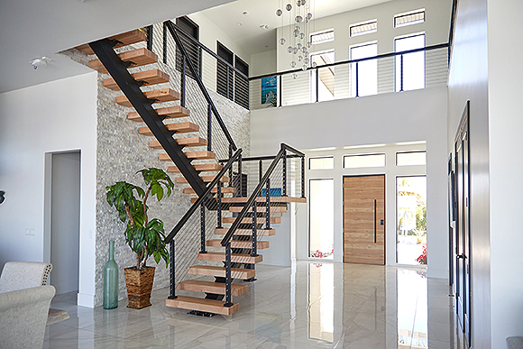 A mostly-white home with a stone wall has an open beechwood staircase with black metal framing and handrails that has a twist and a landing partway up. Light streams in from multiple open windows without curtains or blinds, above and by the front door.