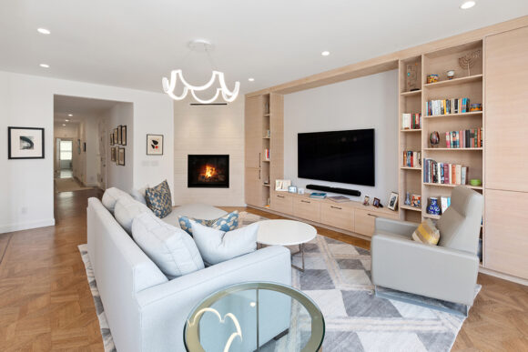 Looking into a living room with an L-shaped sofa, glass side table, white coffee table, and a beautiful geometrically-shaped chandelier above. A fireplace adorns the far wall near the open hallways. On the back wall, there's custom cabinetry in white oak with closed cabinets and single bar metal handles, plus open shelving in narrow widths on either side of the tv opening, and a wider set of shelves to the right. Four cabinet drawers are underneath the television, providing a surface for books, photos, and collectibles. A grey armchair sits in front of the cabinets to the right, with a grey, white, and yellow striped lumbar pillow. A geometrically patterned rug in white, grey, and pale pinkish-tan that matches the cabinetry creates a zone for the furniture.