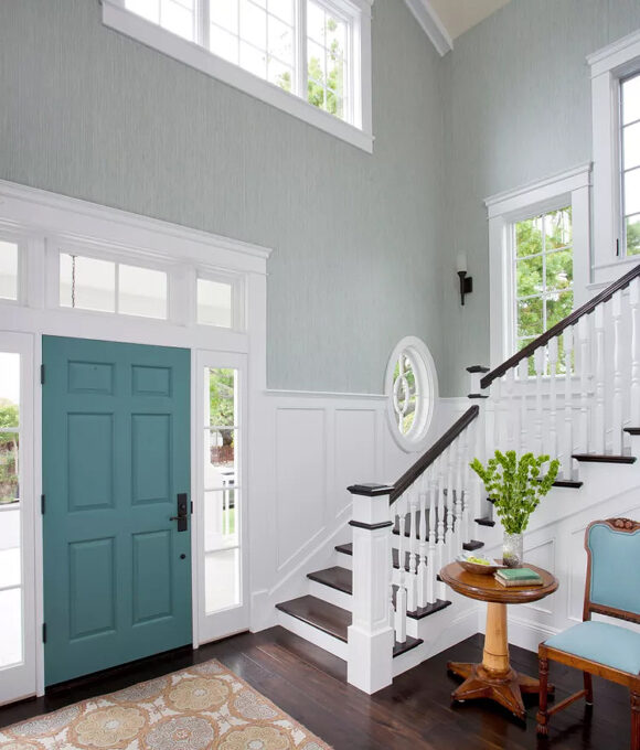 A high-ceilinged entryway with lots of windows wrapping around and going up the stairs to the right in classic white with as dark wood bannister and matching steps. The walls are painted a light seafoam color, and the door is a dark turquoise hue. A round pedestal table in cherry wood holds a plant and books next to a cherry wood chair with sky-blue upholstery.