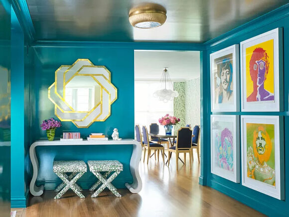 Turquoise paint on the walls and trim envelop you in this entry. A geometric frame shaped mirror is hung above a console table dressed with flowers and books, and two stools for putting shoes off and on are finished in a silver sparkly material. Four colorful artworks of The Beatles take up the entire right entry wall. You can see a dining table and chairs just beyond, through the hallway opening.