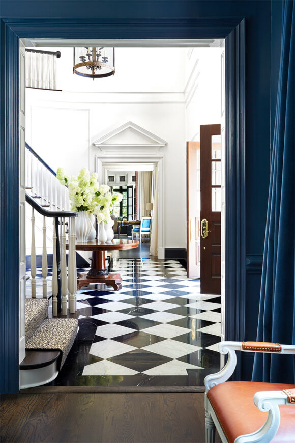 Looking into a classic entryway surrounded by blue paint on the doorframe and walls. Inside the space, black and white checkered flooring stretches to the other room. A black staircase with white balustrades extends upwards to the left. In the center is a cherry wood table supporting multiple white vases of white flowers. Just beyond that is a wide wall opening with a column-type pitched top above the doorway in white to match the walls. A chair, lamp, and desk are visible beyond, with creamy curtains and dark window frames.