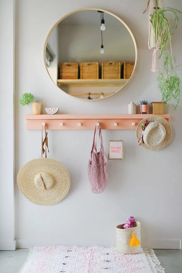 A light-colored entry with a round blond wood mirror reflecting 4 woven storage boxes up on the shelf, and a pair of exposed lightbulbs hanging down, behind the viewer. A peach-colored shelf with 7 silver pegs bearing a pair of straw hats, a net bag in pink, and a sign reading "You Are Magic" hanging from a hook, while several small plants dress the shelf, and a hanging plant's green vines extend down on the right. The flooring is basic grey concrete with a pink and white rug, and a woven basket with a pink umbrella inside awaits a rainy day.