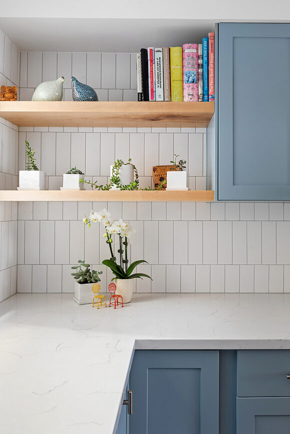 A close-up view of the corner with veined white countertops, vertical white tiling, cornflower blue upper and lower cabinets, warm wood open shelving dressed with plants, cookbooks, and sculptures of birds. On the counter is a pair of flowers in square white pots, with a pair of tiny chairs, one in yellow and one in orange, next to the flowers.