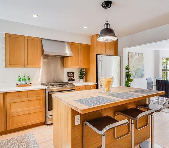 The same kitchen but in warm wood tones, white countertops, and silver appliances. No color other than wood, white, silver and black. 3 modern black and silver counter height stools sit in front of the butcher block island, which is finished entirely in matching wood vertical surfaces to the countertop.