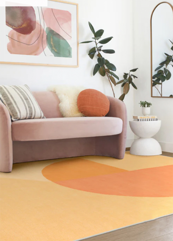 On the floor, an area rug in creamy orange and a 3/4 circle of darker orange. Upon the rug sits a pink loveseat sofa with 3 throw pillows: A grey vertical stripe, a round solid orange, and a white faux fur. Next to the sofa is a white modern side table dressed with two books and a small plant. Next to the table are a wall-hung mirror and a fiddle plant. Above on the wall is a pink and green and maroon abstract artwork.