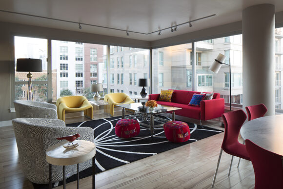 A living room with a double aspect window corner wall and view of city buildings in San Francisco. Inside the living room, there is a red sofa dressed with primary colored pillows of yellow and blue. Nearby are a pair of modern yellow chairs, a pair of grey patterned tub chairs, a glass coffee table, and a pair of red poufs on a black rug with white geometric lines.