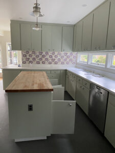 BEFORE kitchen light renovation, with sage green cabinetry, a wooden kitchen island countertop, and white counters with purple flowered wallpaper as backsplash.
