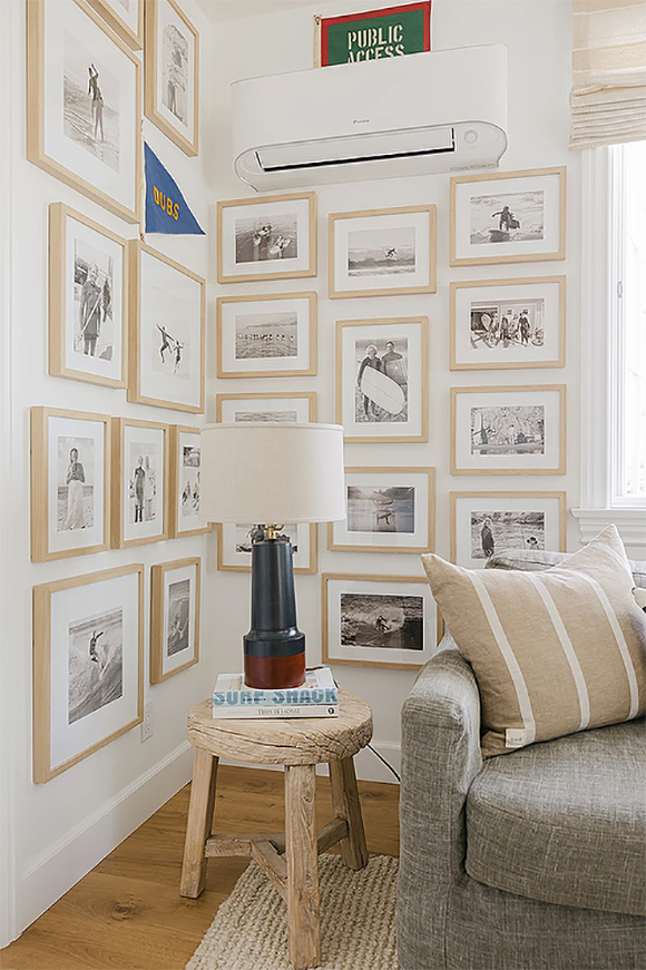 Two photo walls joined in a corner, created with blonde wood frames and black and white images. A stool acts as a side table with a table lamp in black with a white shade sat upon two books. Next to that, the end of a textured grey sofa and a beige throw pillow with white stripes.