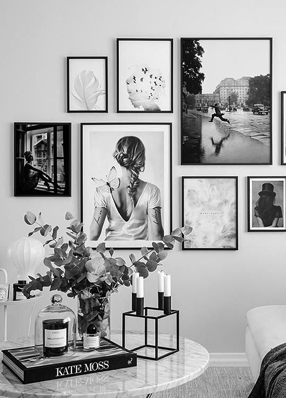 A staggered collection of black and white photos with thin black frames and no matting hung on a white wall behind a marble table dressed with a plant, Kate Moss book, a square candle holder and tapered candles, and a candle under a glass container.