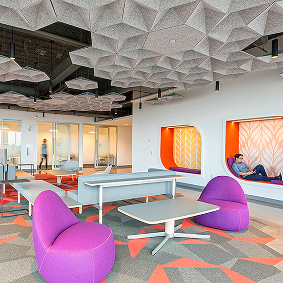 On the ceiling are grey hexagonal shapes that are pushed in to meet in the center, creating a geometric pattern. Where there are no ceiling tiles, a black background shows through, and some lights hang down. 

This is in an open space with purple chairs next to a grey rectangular table with white edging and white pedestal. A matching sofa table is behind an angular grey sofa with more chairs and tables beyond. At the back of the space is a hallway and several glass doors to small meeting rooms. In the wall are two cutouts that allow a person to lay or sit down in and on them. The cutouts are orange with wallpaper on the inside walls.