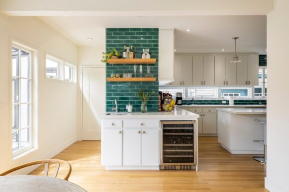 The L-shaped end of the kitchen counter, now with wood open shelves and green Ann Sacks subway tile, plus a wine fridge next to the drinks sinks and white cabinetry below white countertops.