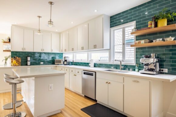 AFTER light renovation, a kitchen with white cabinetry, white countertops, and jewel-green subway tiles, plus stainless steel appliances. Open wood shelves on the right end of the counter near the sink. On the left side of the kitchen, an island with counter seating.