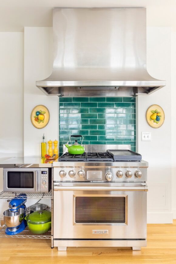 Same stove but updated with green subway tiles that match the rest of the kitchen, plus side shelves to hold the KitchenAid, cookware, and a shelf for the microwave, with a metal countertop above for cooking that creates some open space next to the cooktop. A bright green teapot sits on the hob.