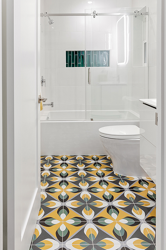 Colorful geometric shapes in cornflower yellow, muted blue-green, black and white are the standout feature of this white bathroom. A shower niche inset with green tones with the flooring to make it feel coordinated.