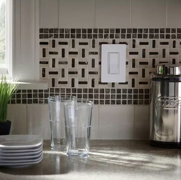 A white Lutron rocker light switch with a dimmer slider to the right side, on a wall over a tiled backsplash in a kitchen, above the counter with glasses, a stack of white plates, and a coffee canister in silver and black. A tiny peek of a window and a green plant are just visible to the left.