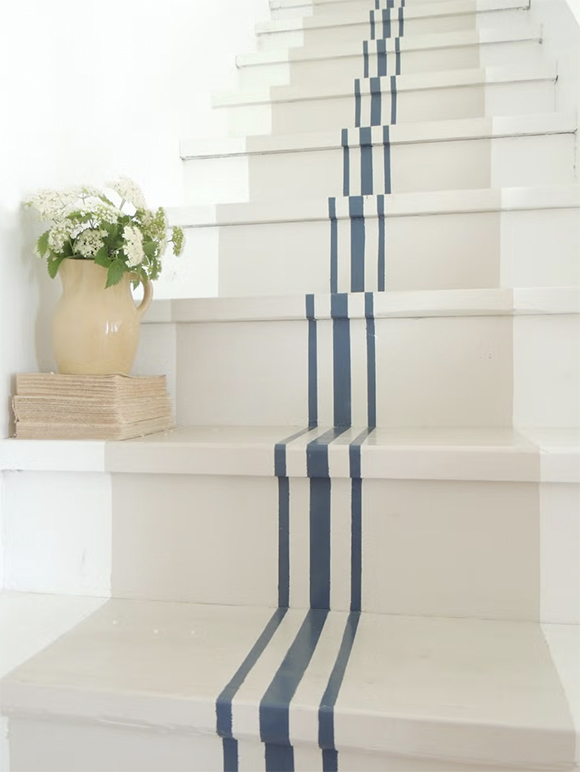 A beautiful white painted staircase with beige center portion accented with 3 country-blue colored stripes up the middle, the center stripe being the biggest. A beige jug filled with green and white flowers sits atop a stack of books on the second step.