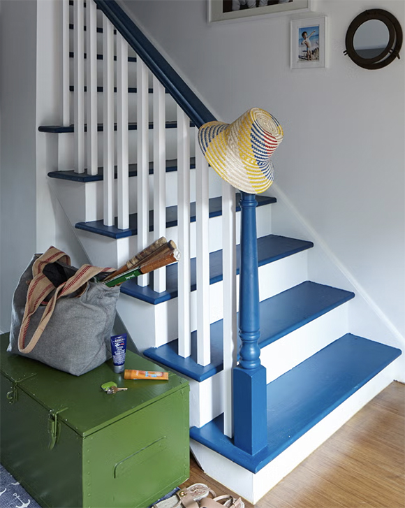 Blue painted stairs with white railings and stringers with a hat on the newel post. A green trunk sits nearby with personal items on it, and a small mirror and art hang on the white wall going up the stairs. Warm wood flooring peeks out at the bottom of the staircase.