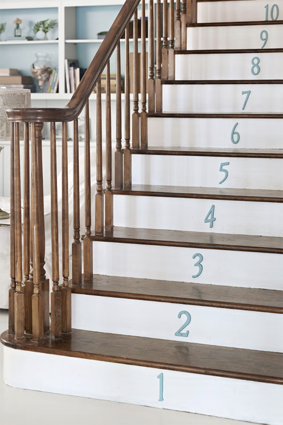 A staircase with dark wood treads and risers painted white, with aqua blue numbers from 1 to 10 on the risers going upwards. In the background, shelves filled with vases, books, and collectibles are slightly out of focus to the left of the stairs, behind the railings that ends in a circle of rails at the bottom step.
