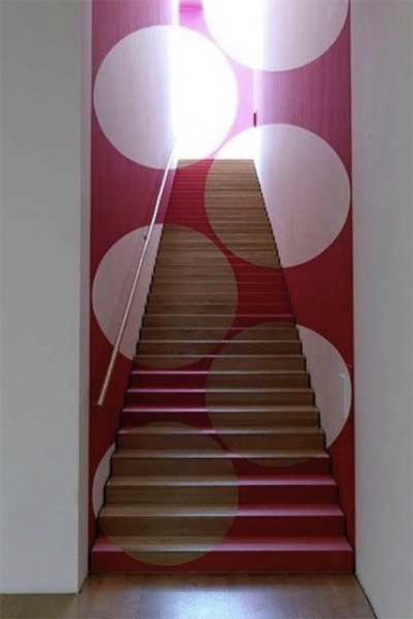An optical illusion of polka dots floating in space is created by painting stairs and their surrounding walls with pink and white paint, including the door at the top of the stairs, and also revealing the wood color for some portions of the dots. Light beams from around the doorway.