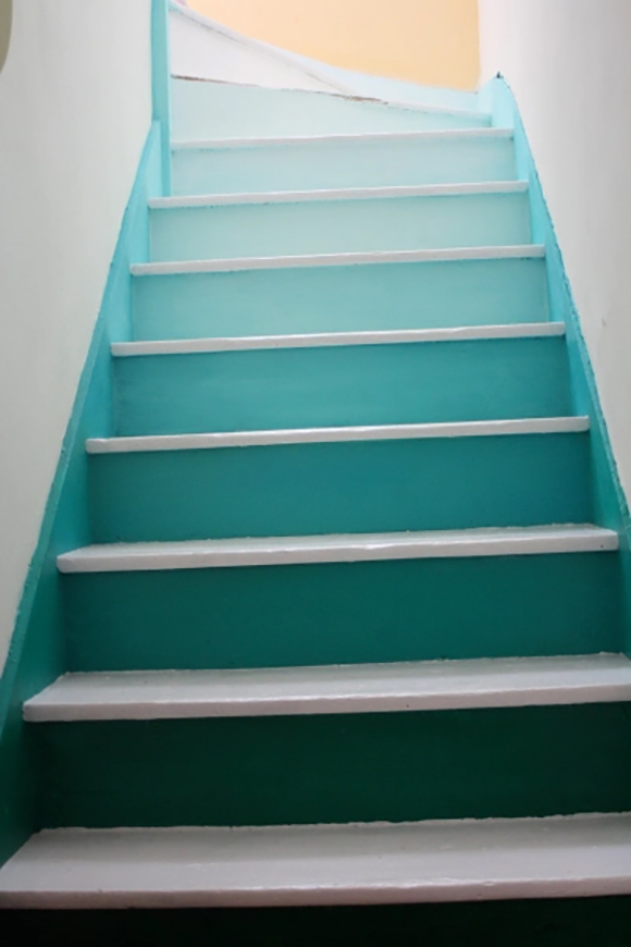 A staircase at a slight angle in the photo starts with dark treads at the bottom, gradually lightening the colors through shades of blue and turquoise, rising to a pale blue at the top where the stair turns to the left. This graduated change in color is called ombré. The treads themselves are all painted white, as are the surrounding walls. The stringers are painted a solid turquoise blue to harmonize the colors.