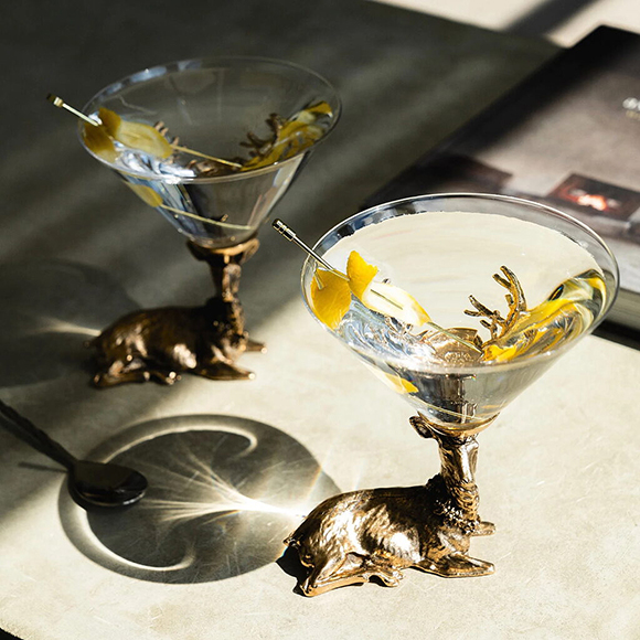 Two custom stemware glasses formed from kneeling stags with the drinking glass balanced between their antlers are displayed on a tabletop. Light shines through the first glass, revealing a gold pin holding lemon slices inside the clear drink. The second stag glass is behind in shadow and slightly out of focus.
