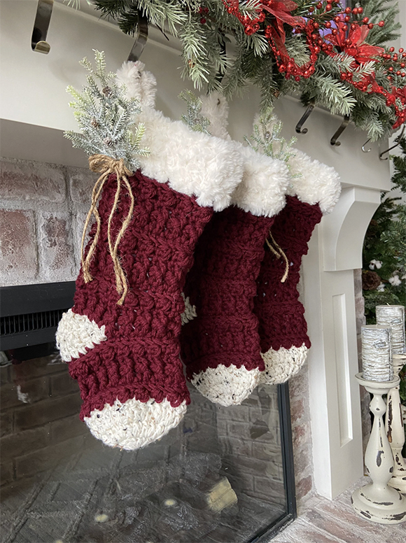 3 hand-crocheted red and white stockings hang over a fireplace with greenery tied to the white cuffs with packaging string for an old-fashioned look. Two white and grey candles stand to the side of the fireplace surround, which is painted white, while red berries and green pine needles hang down from the mantel above. These stockings are examples of Nonna Holiday style designs.