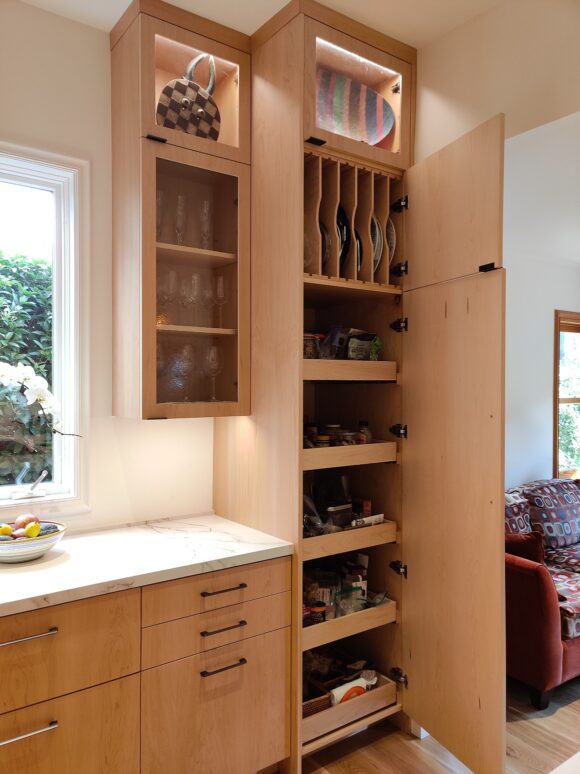 A floor-to-ceiling pantry with pullout drawers and plate storage hides behind light oak custom cabinetry. A lighted glass cabinet at the top allows for displaying without having to dust or clean greasy residue.