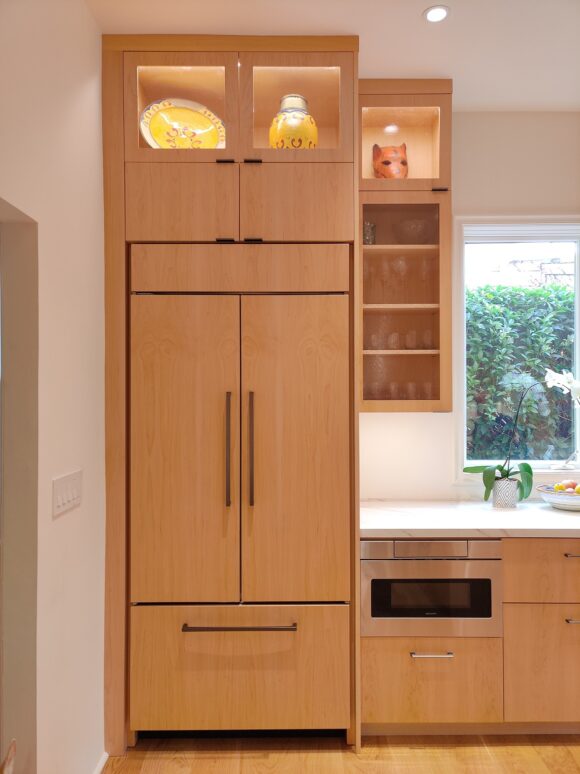 A SubZero fridge is concealed behind a panelled custom oak cabinet with double doors on top and a single freezer drawer below. To the left is an under counter microwave drawer in stainless steel. Above are additional cabinets to the ceiling. At the top is a glass-fronted cabinet with built-in lighting for displaying collectibles and kitchenware. The next glass cabinet is for drinking glasses. A window outlined in white allows natural light to stream into the kitchen, bouncing off the white sinterset quartzite countertops and matching backsplash.