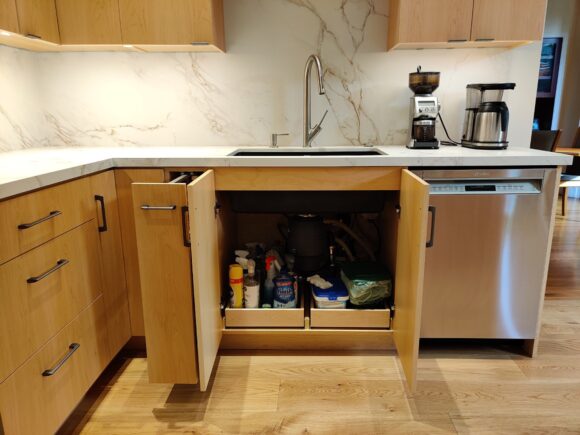 Double doors under the kitchen sink reveal two pull-out trays for cleaning supplies, keeping everything neat and accessible. To the right is a stainless steel dishwasher. To the left is a slide-out spice rack. The veining is visible on the backsplash made of sinterset marble-look quartzite. Much better than real marble because it doesn't stain or damage easily.