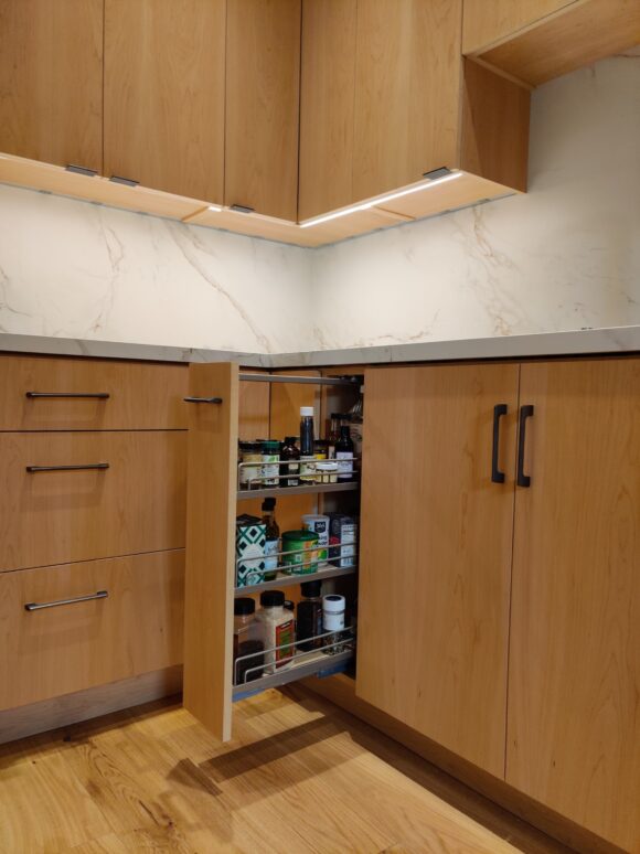 A custom slide-out spice rack sits next to the sink and near the range for easy access. The cabinet front is in the matching light oak finish as the rest of this custom kitchen. Under cabinet task strip lighting is visible above, along with a row of convenient electrical outlet plugs.