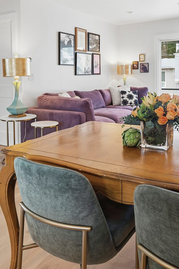 Looking from behind the green dining table chairs and warm wood table dressed with orange and green flowers and an artichoke, we can see the living room just beyond, with the purple couch, framed photos, side tables, and lamps.