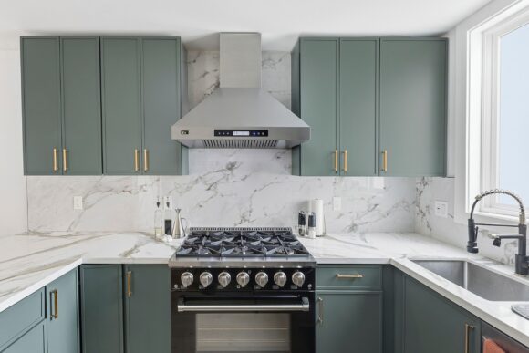 A head-on look at the kitchen, with upper and lower cabinets in moss green and gold hardware, contrasting the white marble-look counters and backsplash. A black and gold faucet hangs over the silver undermount sink to the right side. A black with silver range and silver hood above sit in the center. A window is peeking off to the right behind the sink.