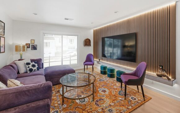 Living room with white walls and ceiling, purple sofa with matching footrest, orange patterned rug, 2 purple chairs facing the sofa, a glass coffee table, and 3 jewel-green poufs tucked underneath a slatted wooden wall with under-mount lighting that shows off the large television. Pillows, bird sculptures, artwork, and tableside lamps provide interest. The floor is a medium warm wood color. Light streams in through the double window, while recessed lighting is also provided above.