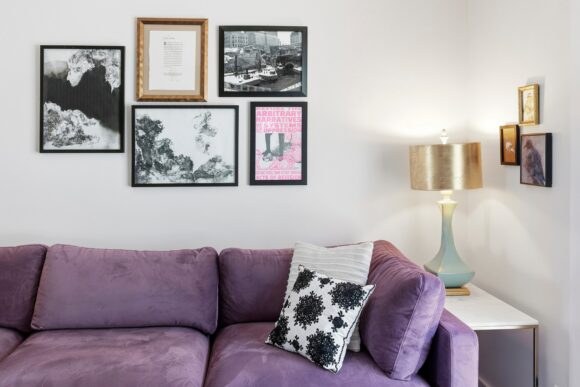 A close-up view of the corner with the purple sofa and throw pillows, 5 framed photos on the wall behind, one of which is pink and the rest are black and white, with 3 more small artworks on the 90-degree wall. A light green ceramic lamp with a gold lampshade sits on the narrow white side table, casting light and shadows.