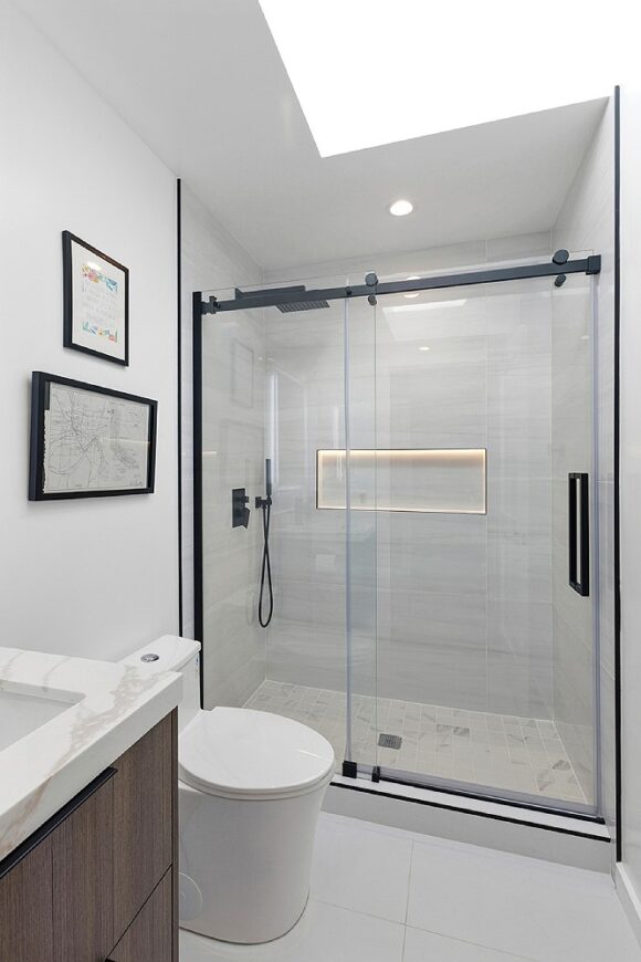 A mostly-white bathroom with black hardware. A skylight above provides light in addition to the recessed lighting above the shower, which is glass with black framing and has black shower handle and knobs. A lighted recessed shower niche is filled with off-white tiles that match the rest of the shower, while the off-white and grey floor tiles are coordinating but not matching. A white toilet sits between the shower and the sink, which is finished with dark wood cabinetry and marble-look counters. Two artworks are framed in black on the wall above the toilet.