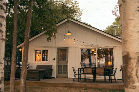 Exterior of a white-sided backyard building or ADU with string lights, outdoor seating and dining, and a BBQ.